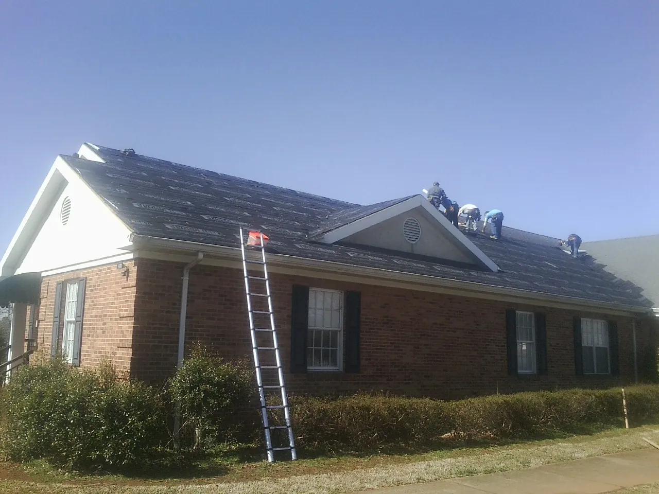 Crew tearing shingles off a brick ranch roof with a ladder set — Metal Roof Repair work by our crew in China Grove